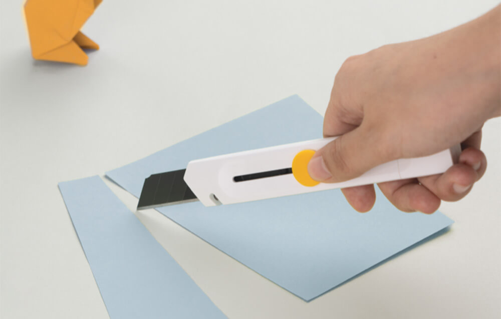 Hoto Cutter für 18 mm Klingen Mit einem Hoto Cutter für 18 mm Klingen wird ein Stück hellblauer Fotokarton zerschnitten.