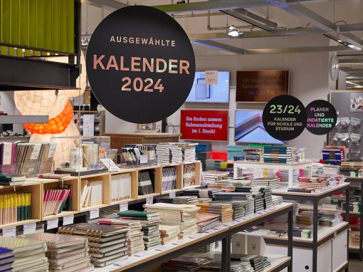 Im Modulor-Laden liegen auf Tischen viele verschiedene gebundene Kalender und Planer in Stapeln nebeneinander. Darüber hängen kreisrunde Schilder in schwarz mit goldener Aufschrift "Ausgewählte Kalender 2024" sowie "23/24 Kalender füd Schule und Studium" sowie "Planer und undatierte Kalender"