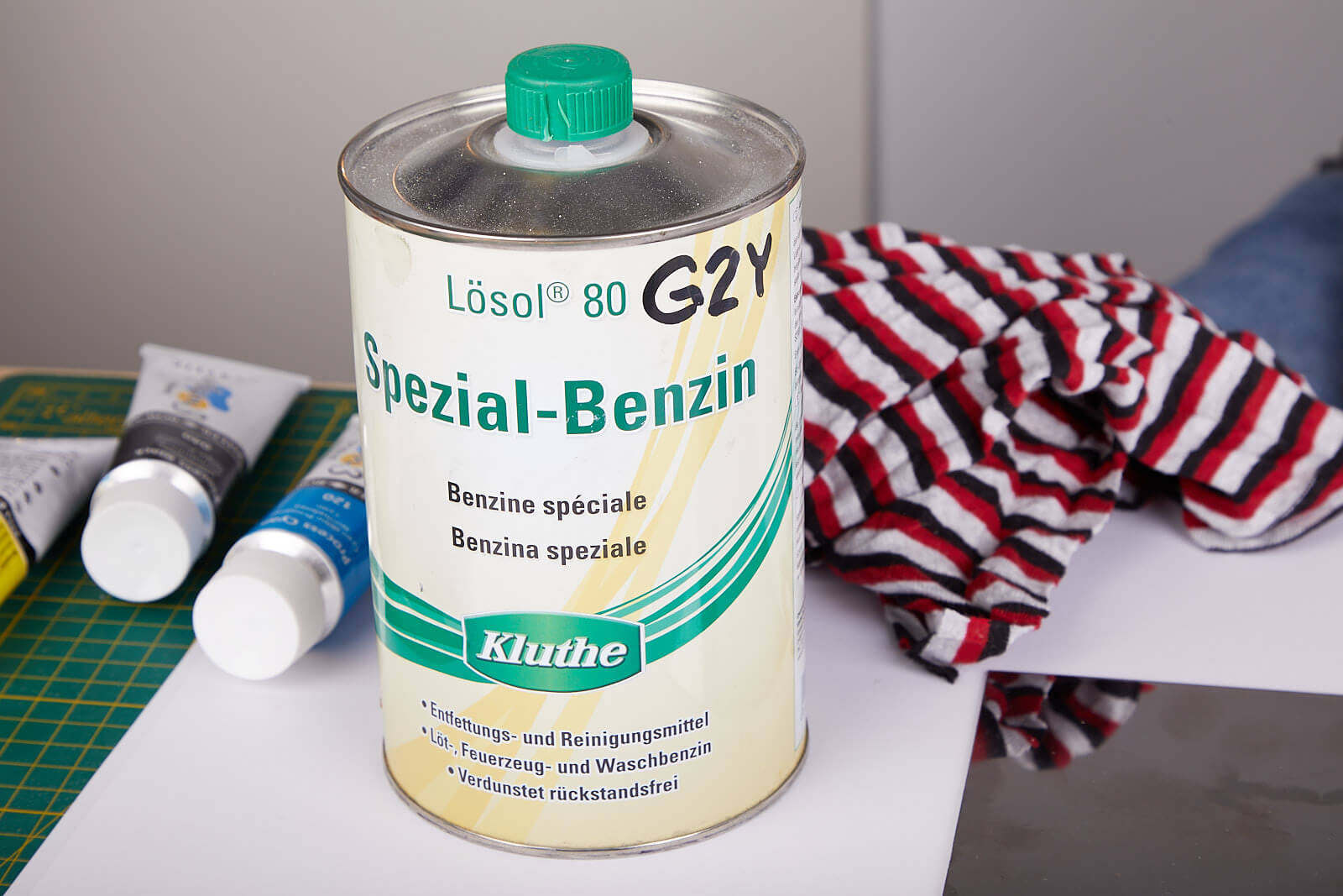 Lösol Spezial-Benzin eine Blechflasche (1 Liter) Spezial-Benzin zum Entfetten steht auf einem Tisch. Daneben liegen ein Lappen und zwei Acrylfartuben.
