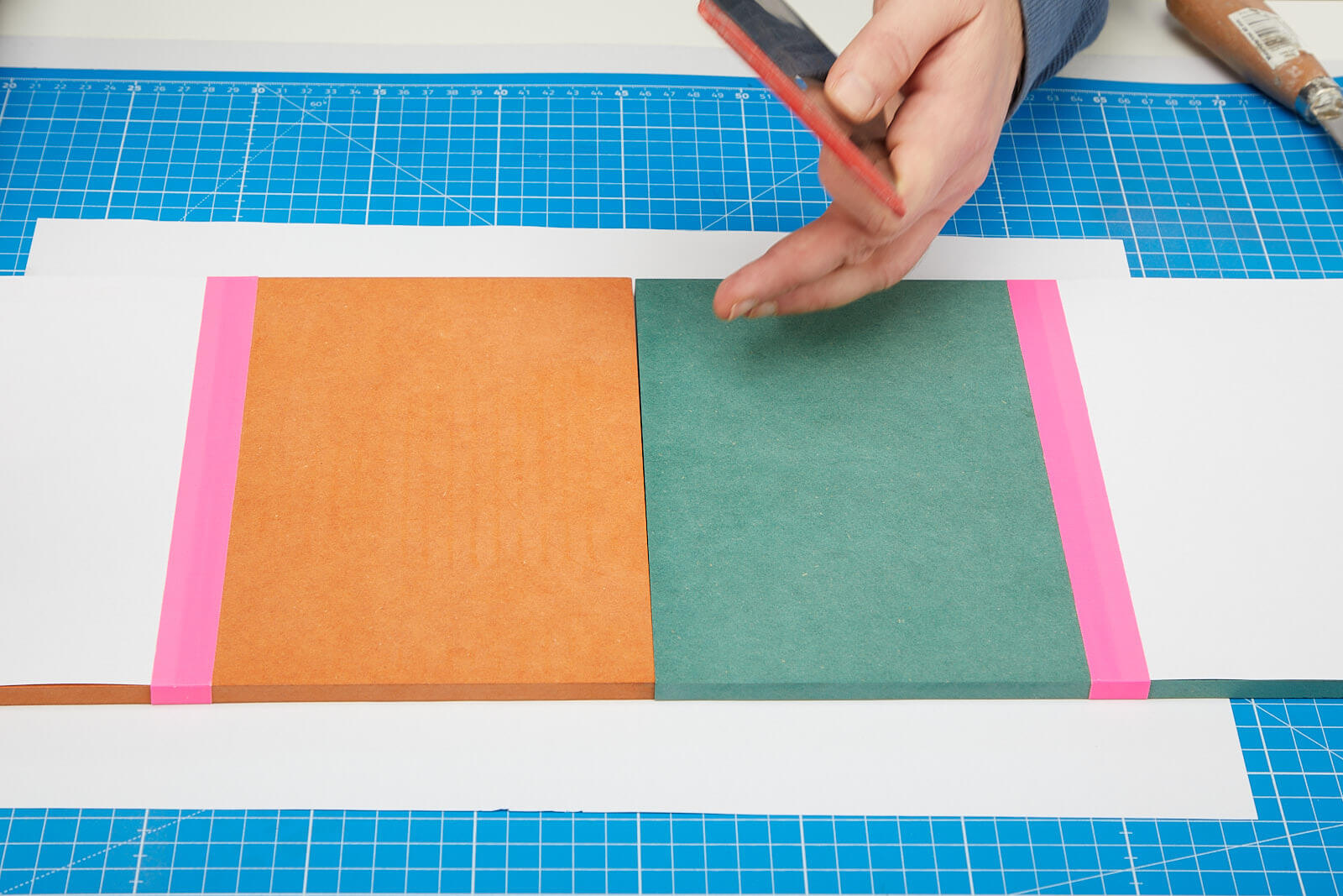 Auf einer Schneidematte liegen zwei Platten farbiges MDF. Die Platten sind zur Hälfte jeweils mit einem Papier abgedeckt. Die sichtbaren Bereiche sollen mit Öl bestrichen werden.