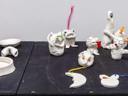 auf einer Unterlage aus schwarzem MDF stehen zahlreiche, von Hand modellierte kleine "Monster" aus weißer Modelliermasse.