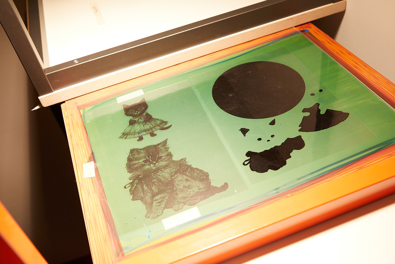 Belichtung Sieb Ein mit Fotoemulsion beschichteter Siebdruckrahmen wurde mit Motiven für das Belichten präpariert; Die Motive (schwarze Katzen) sind auf transparenten Folien auf das beschichtete Siebdruckgewebe positioniert. Mit einem Halogenstrahler wir die lichtempfindliche Fotoemulsion belichtet.