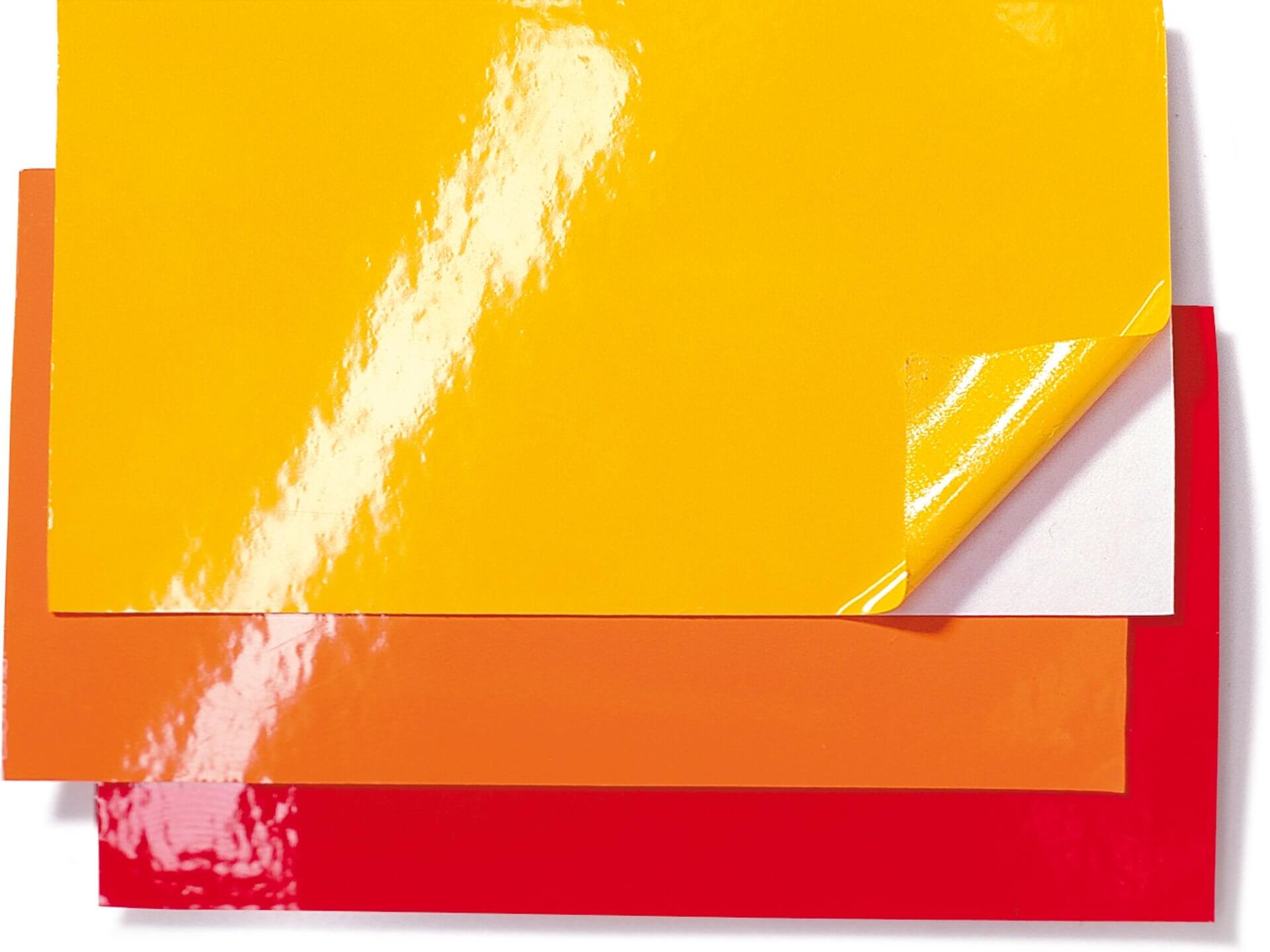 drei kleine Stücke farbige Klebefolie liegen aufeinander. Auf rot liegt orange un darauf liegt gelb. Bei dem gelben Stück ist die Ecke der Folie von der Schutzfolie gelöst und hochgebogen. Auf den flänzenden Folien ist ein langgestreckter Lichtreflex zu sehen (weiß).