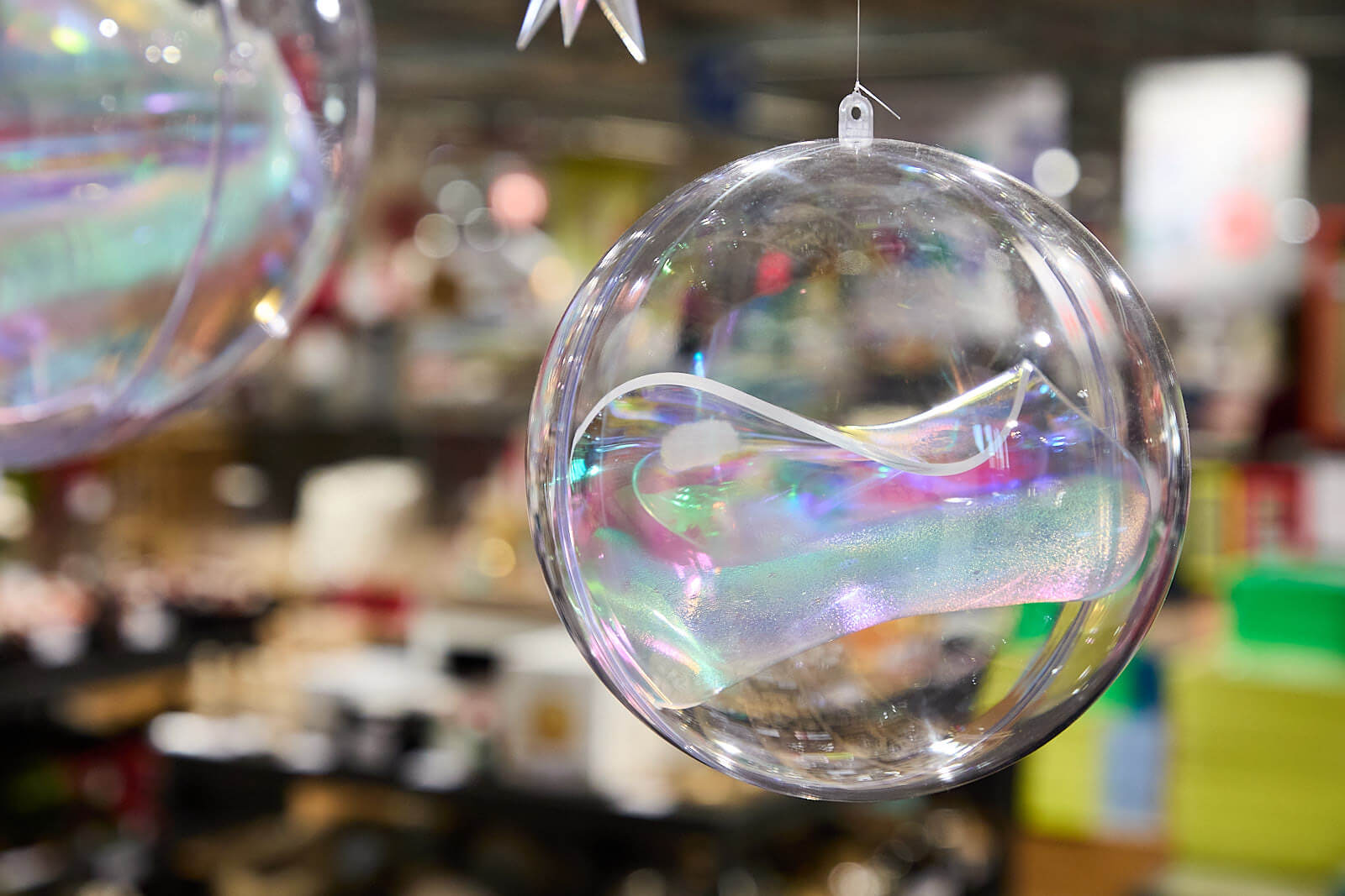 hängende Polystyrol-Kugeln In einer transparenten Polystyrol-Kugel ist eine thermische verformte Acrylglasscheibe aus irisierendem Acrylglas. Die Kugel ist teil einer Ladendekoration, daneben hängt (im Anschnitt zu sehen) eine größere Polystyrol-Kugel. Die Kugeln sind scharf zu sehen, der Hintergrund des Ladens ist unscharf.