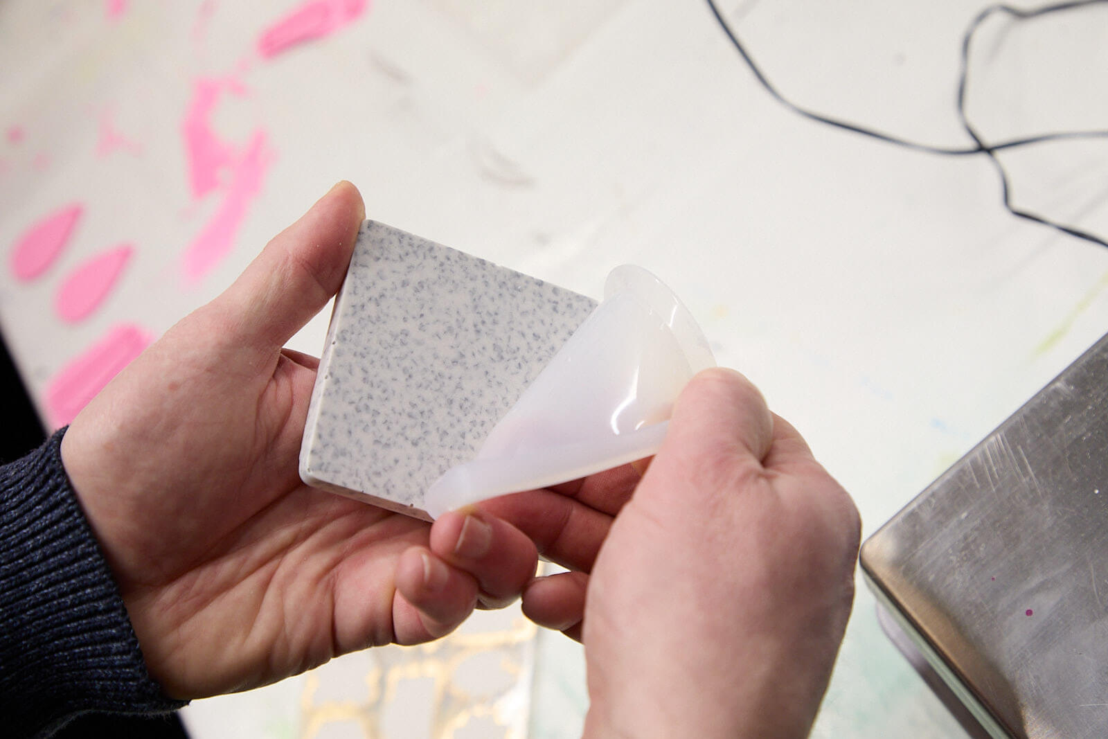 AUs einer transluzenten Silikonform entnehmen zwei Hände eine aus Jesmonite gegossene Fliese. Die Fliese ist mit GlasRoxx-Glaspartikeln versetzt.