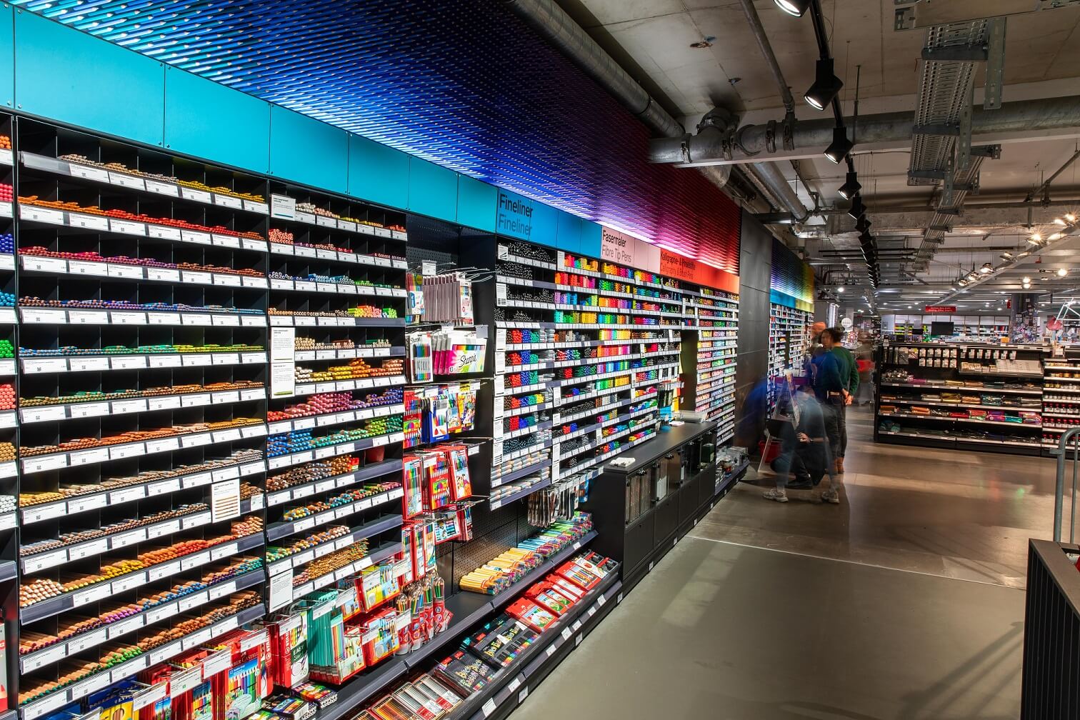 Stiftewand komplett Die Stiftewand wird zu einem großen Teil gezeigt. Im Bild sind hunderte Stiftefächer, deren Regalböden fluchtpunktartig in der Ferne zusammenlaufen. Über den Regalfächern ist die beleuchtete Stifteinstallation zu sehen. In einem Farbverlauf sind Buntstifte in die Wand gesteckt.